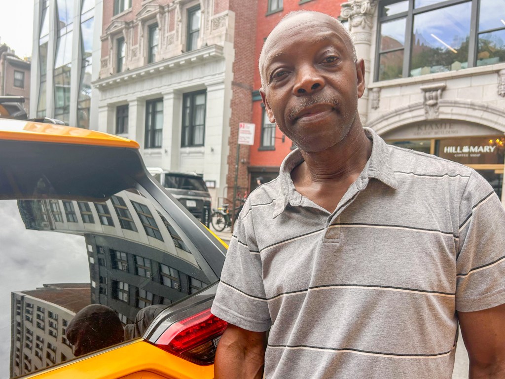 Rafael Gonzaque stands in front of his cab in a grey collared shirt with thin black and white stripes. He has a salt and pepper mustache and looks directly at the camera.