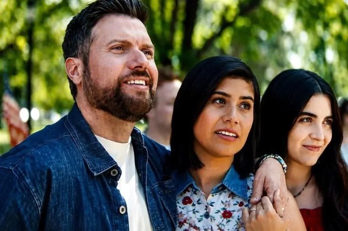 A bearded man in a denim jacket with his arm around a woman in a floral blouse . They, and a young woman with long dark hair next to them, look off camera and smile at an outdoor gathering.