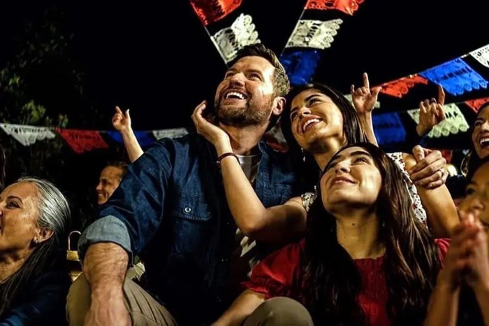 A bearded man and a group of women look up and cheer beneath strings of papel picado at a nighttime celebration.