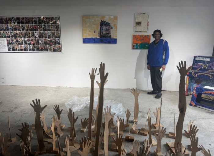 man poses in art gallery with orange painting and sculpture of hands on floor