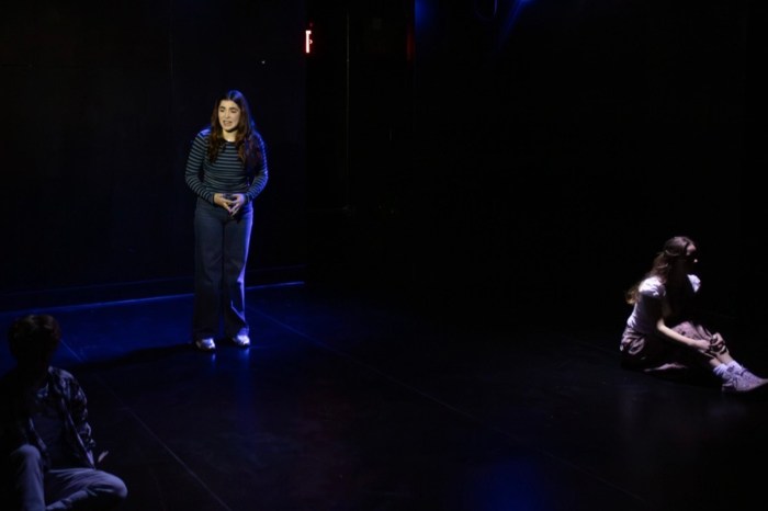 A young woman in a striped top stands alone in blue light on a dark stage while another performer sits on the floor nearby.