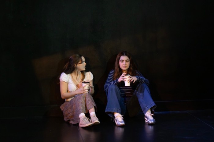 Two young women sit against a wall on a dark stage, holding coffee cups and talking.
