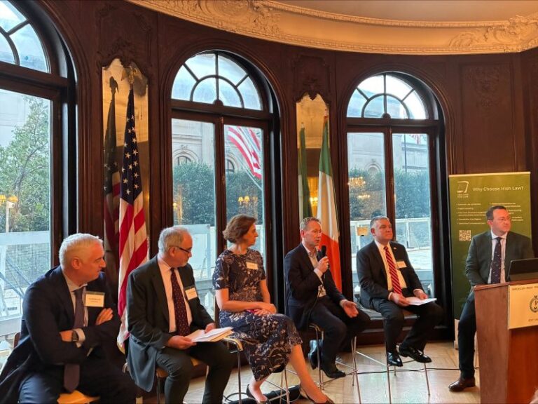 Five business professionals sit and speak on a panel in an elegant room with large windows, flags, and ornate wood décor. One man holds a microphone while others listen. A sign and podium are visible on the right.