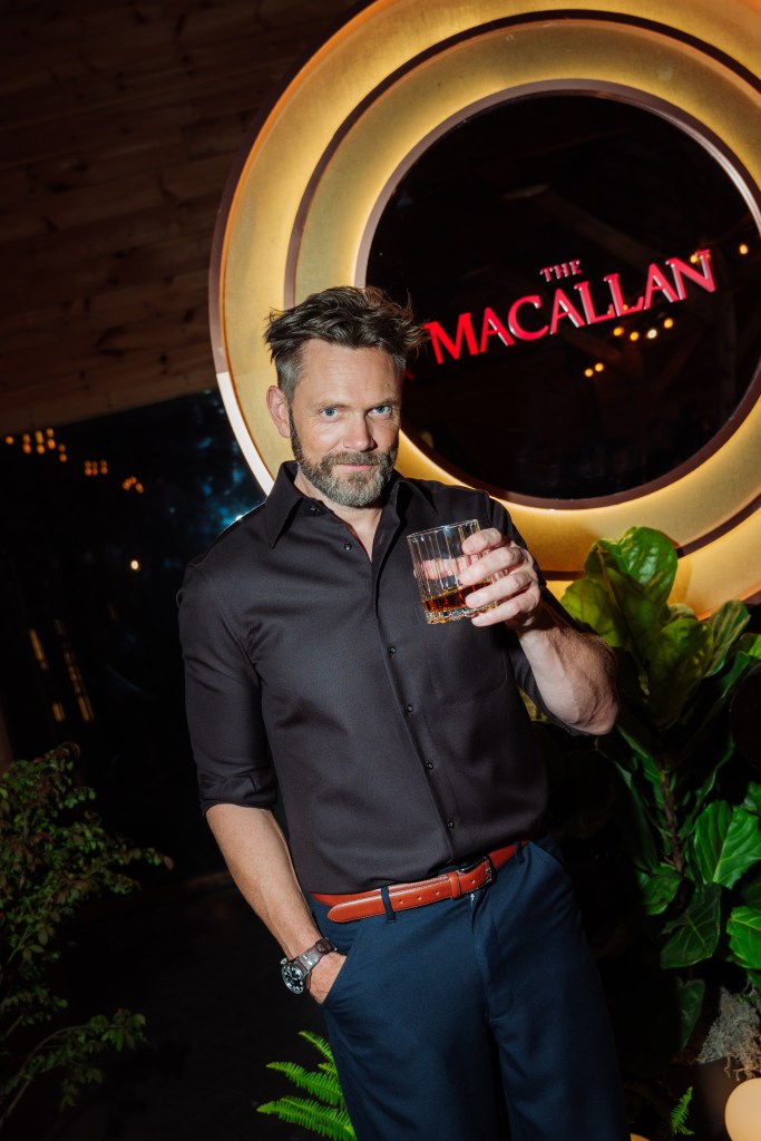 Joel McHale with a drink, smiling in front of a circular sign for The Macallan.