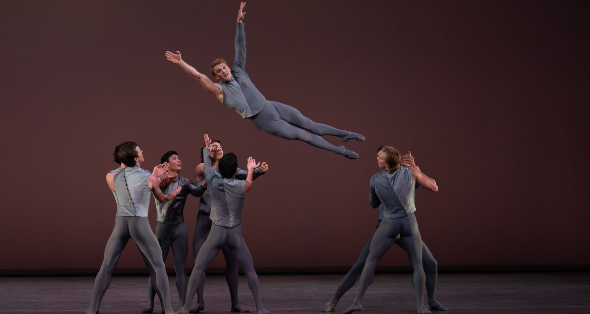 A group of eight male dancers are shown mid-lift onstage. One dancer is midair above the heads of the others, facing the audience with his legs straight and arms in a V above is head, while the rest of the dancers prepare to catch him. All wear medium grey tights and a grey two-toned top that is long-sleeved on one arm and sleeveless on the other.