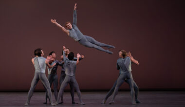 A group of eight male dancers are shown mid-lift onstage. One dancer is midair above the heads of the others, facing the audience with his legs straight and arms in a V above is head, while the rest of the dancers prepare to catch him. All wear medium grey tights and a grey two-toned top that is long-sleeved on one arm and sleeveless on the other.