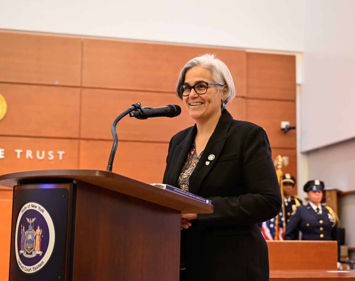 A person with short gray hair and glasses stands at a wooden podium, smiling while speaking into a microphone in a courtroom setting.