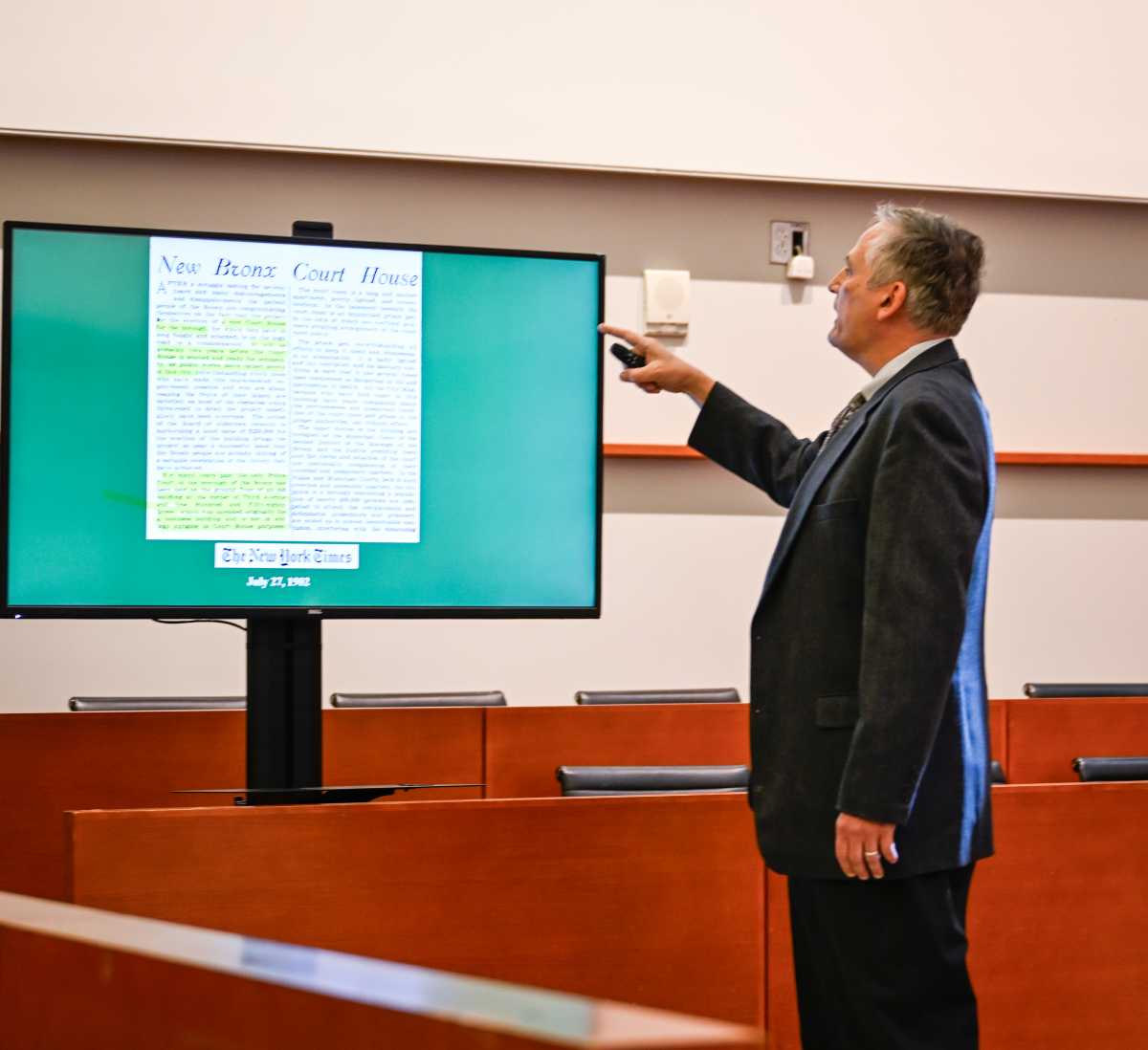 A person in a suit stands in a courtroom, pointing to a large screen displaying a newspaper article from The New York Times dated July 22, 1982, titled "New Bronx Court House."