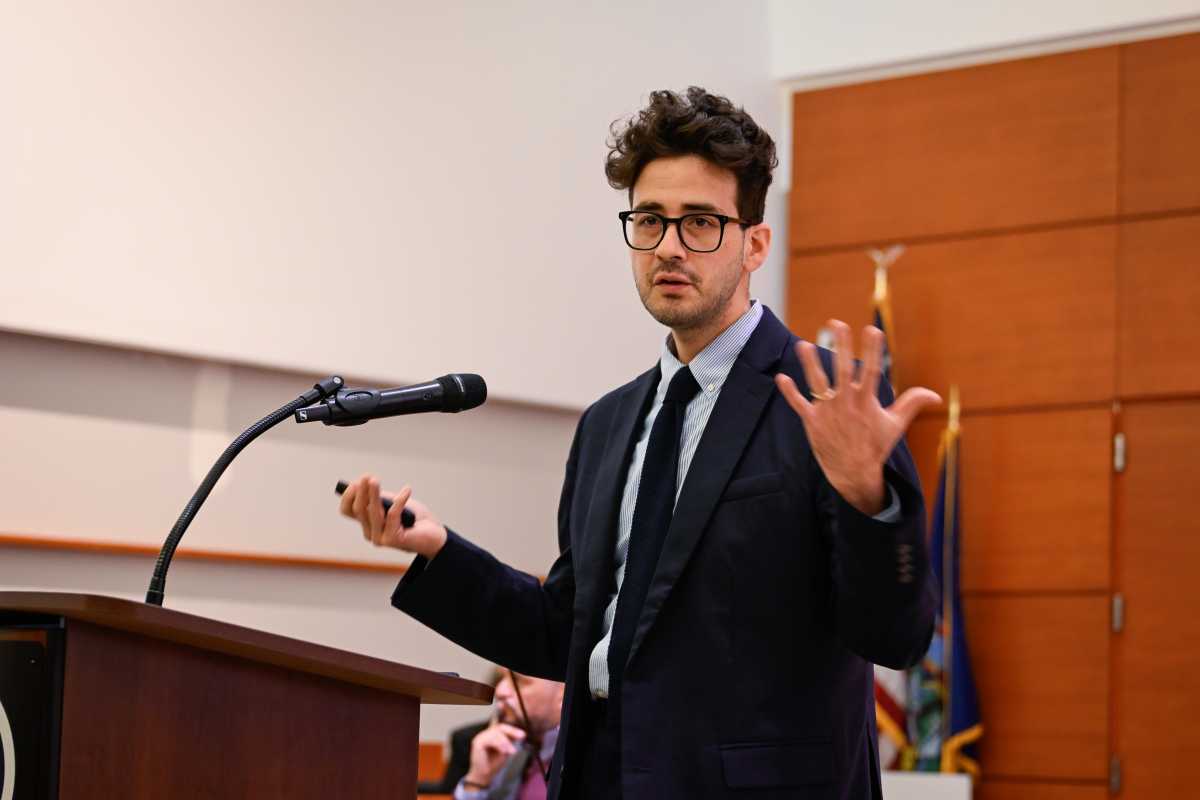 A person in a suit and glasses stands at a podium, gesturing while speaking into a microphone.