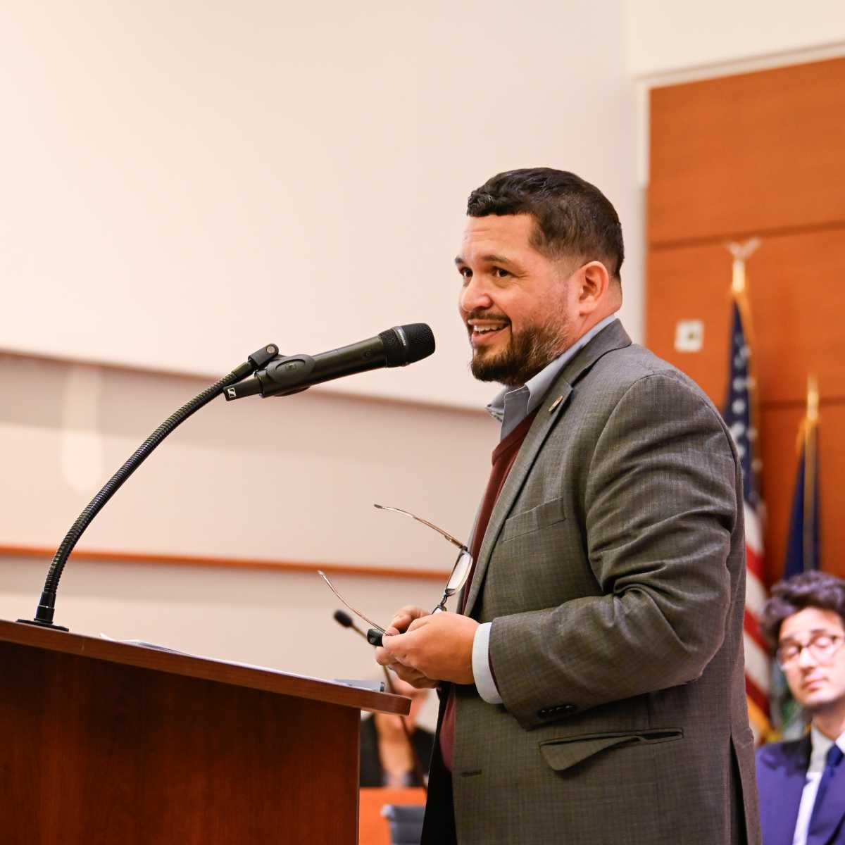 A person in a suit stands at a podium, speaking into a microphone while holding glasses.