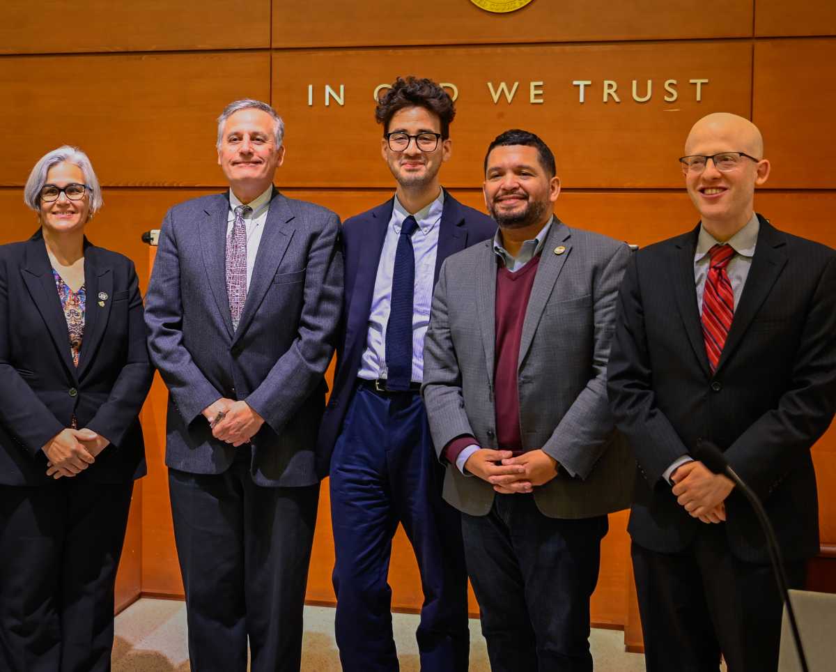 Five individuals in formal attire stand together in front of a wooden wall bearing the inscription "IN GOD WE TRUST."