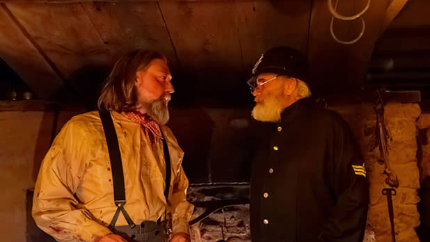 Two bearded men in rustic historical clothing converse inside a dim wooden cabin, lit by amber light; one wears a uniform and cap during the Murder, Monsters, & Mayhem Dinner Theater at Genesee Country Village & Museum.