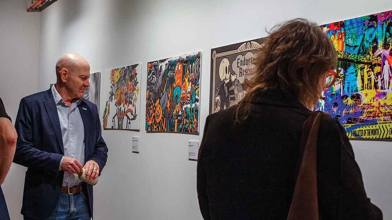 Two people look at colorful paintings hung on a white wall.