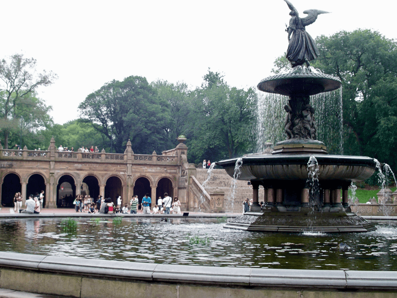 A fountain with an angel statue in the center, surrounded by a pond and green trees, located in a park with people walking around.