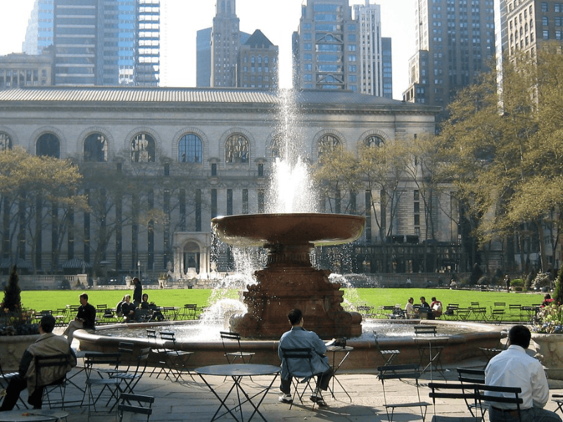 A view of a fountain in a public park surrounded by tall buildings, with people sitting at metal chairs and tables, enjoying the space.