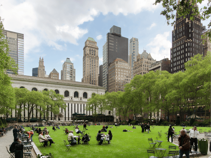 A bustling urban park scene with people relaxing on a grassy area surrounded by tall city buildings, including a historic library, under a partly cloudy sky.