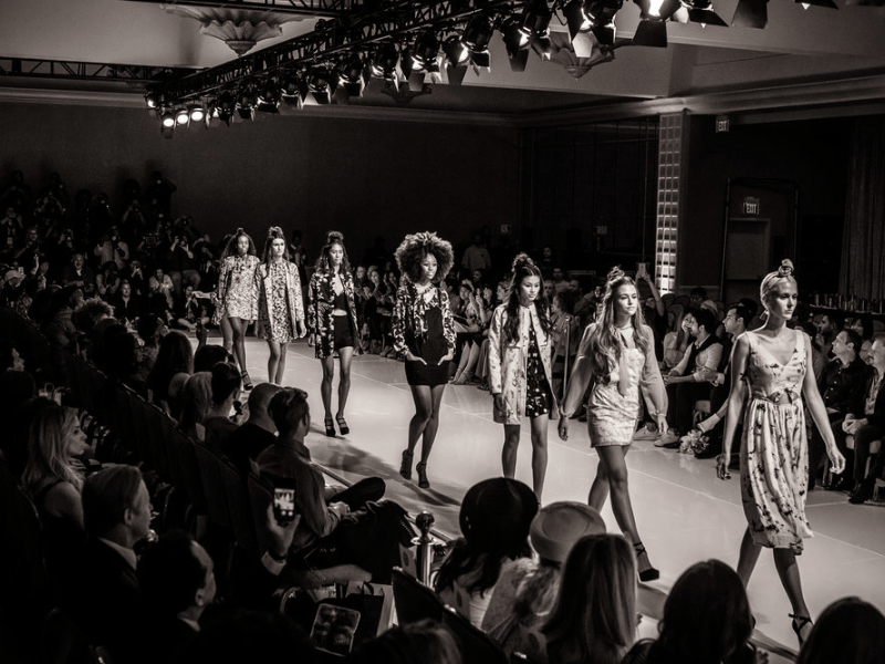 Black and white image of a fashion runway show featuring multiple models walking down the catwalk, with an audience seated on either side.
