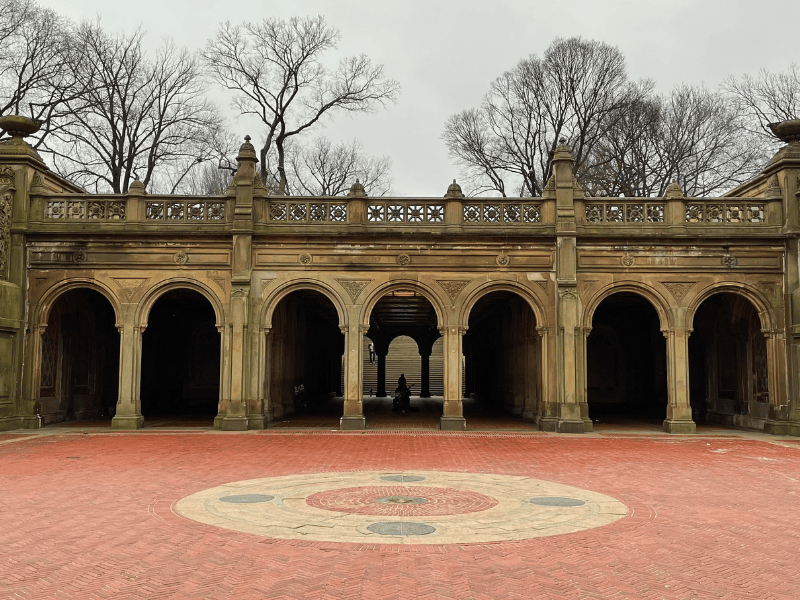 A stone archway structure with multiple arches and decorative details, leading into a walkway with a circular brick pattern in the foreground, under a cloudy gray sky.