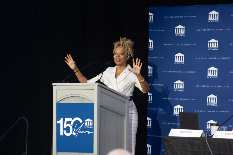 A woman in a white outfit speaks at a podium with 150 Years on it, gesturing with raised hands. A blue NYSBA backdrop and a panel table with nameplates are in the background.