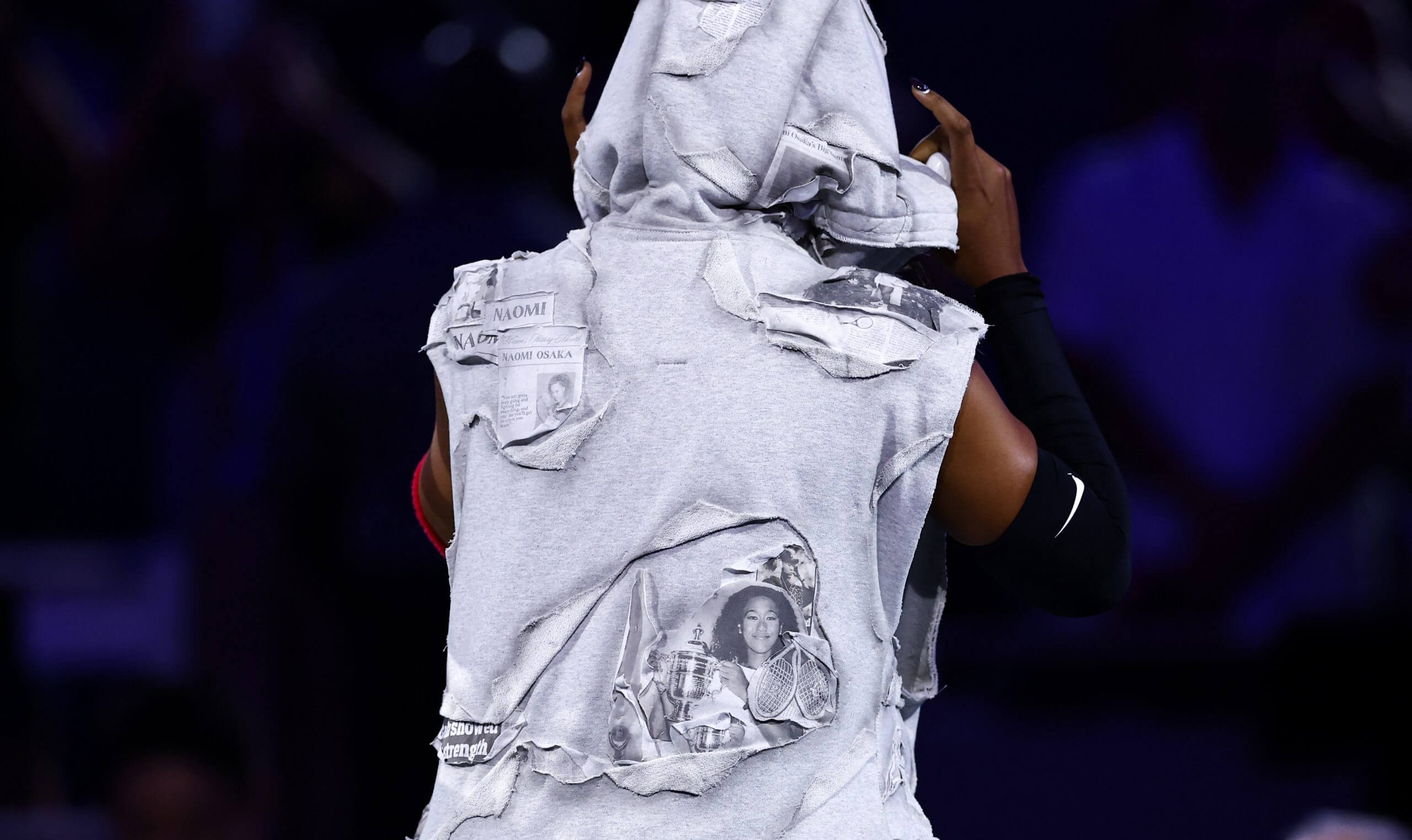 Naomi Osaka faces away from the camera wearing a French terry hooded sweater with newspaper clippings stitched into it.