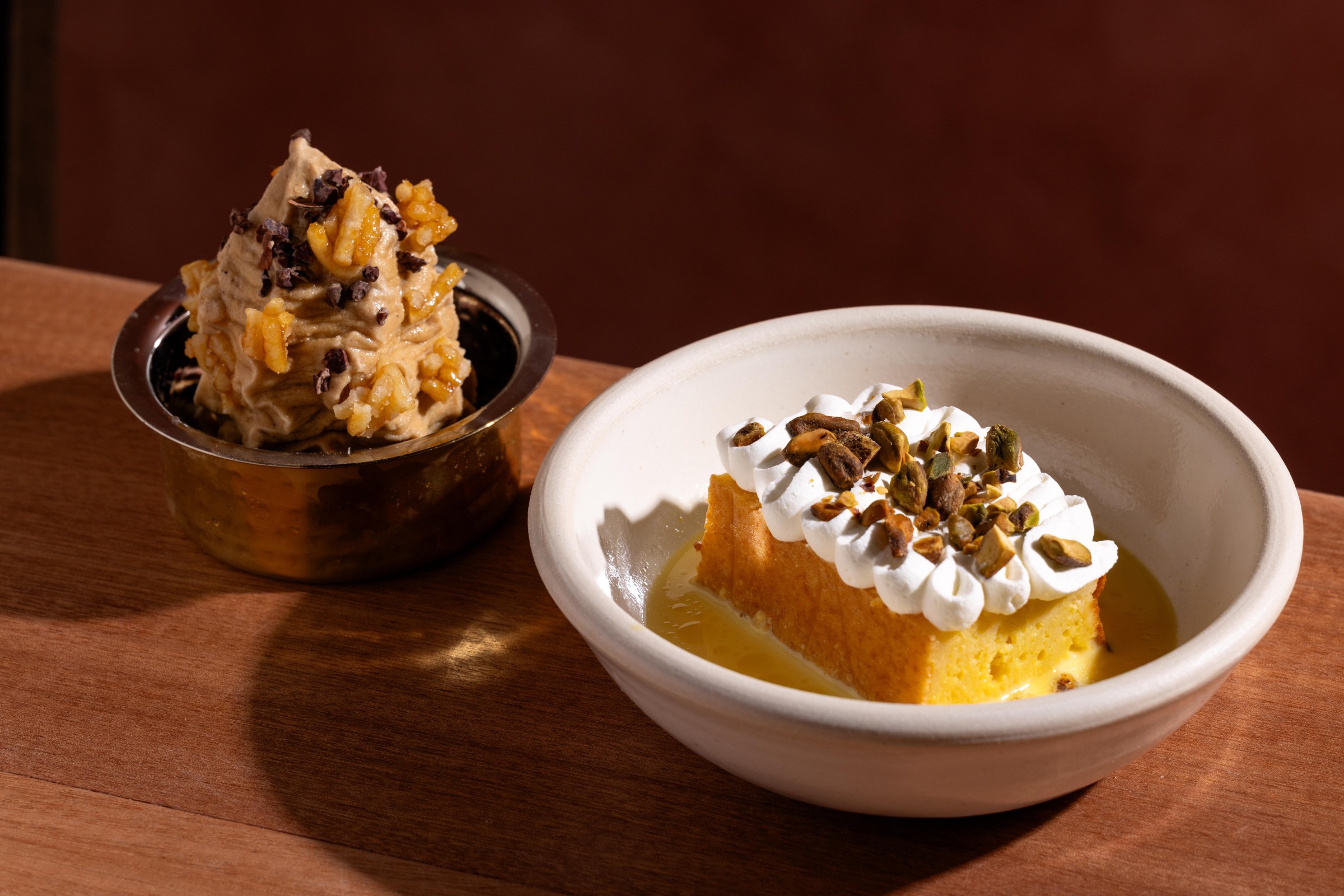 A metal bowl of brown ice cream next to a white bowl of a cake.