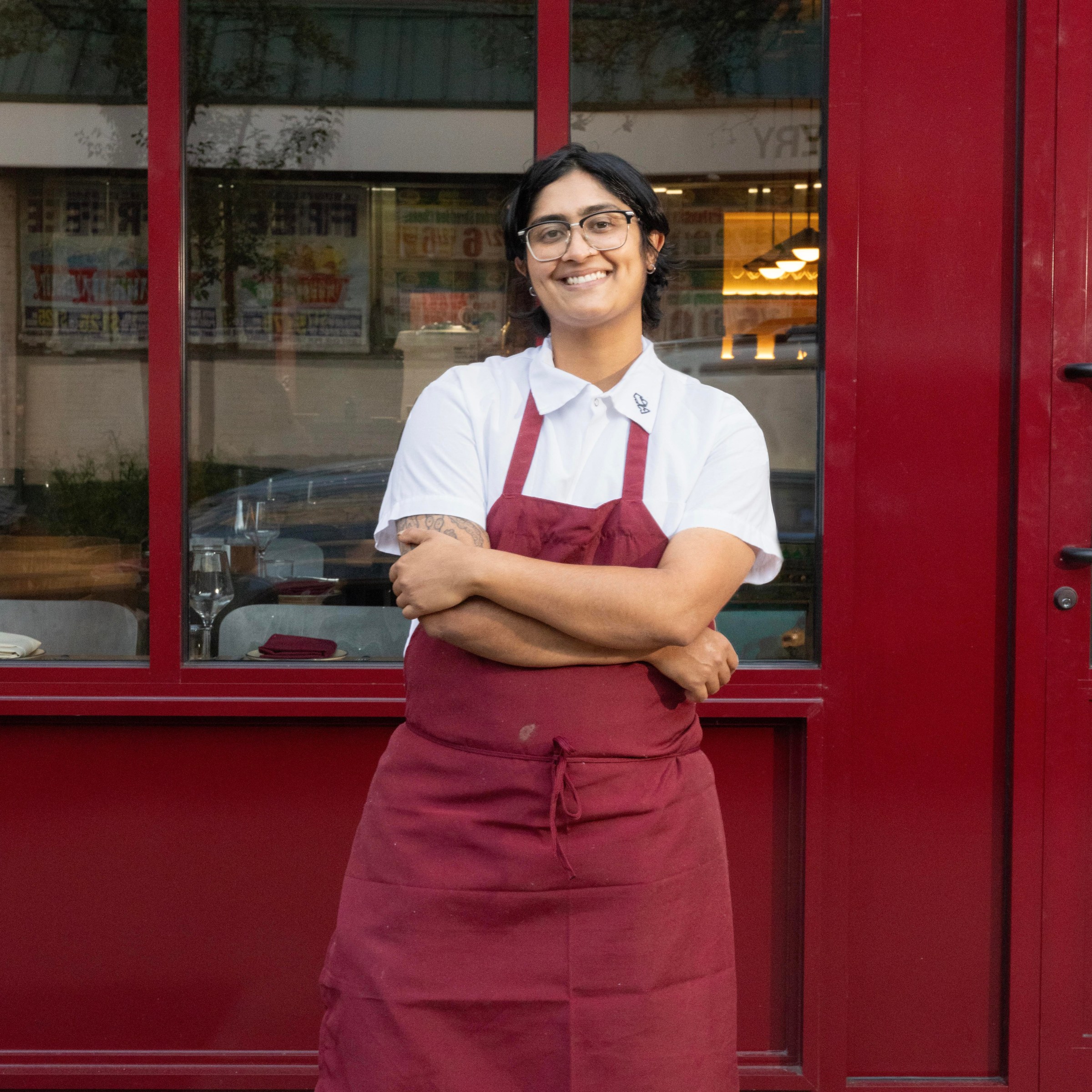 A person in a white shirt and red apron in front of a red building.