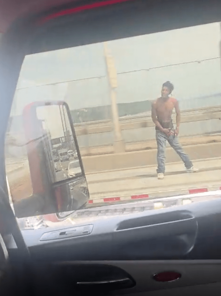 A shirtless man in jeans and white sneakers on a bridge, viewed through a car window.