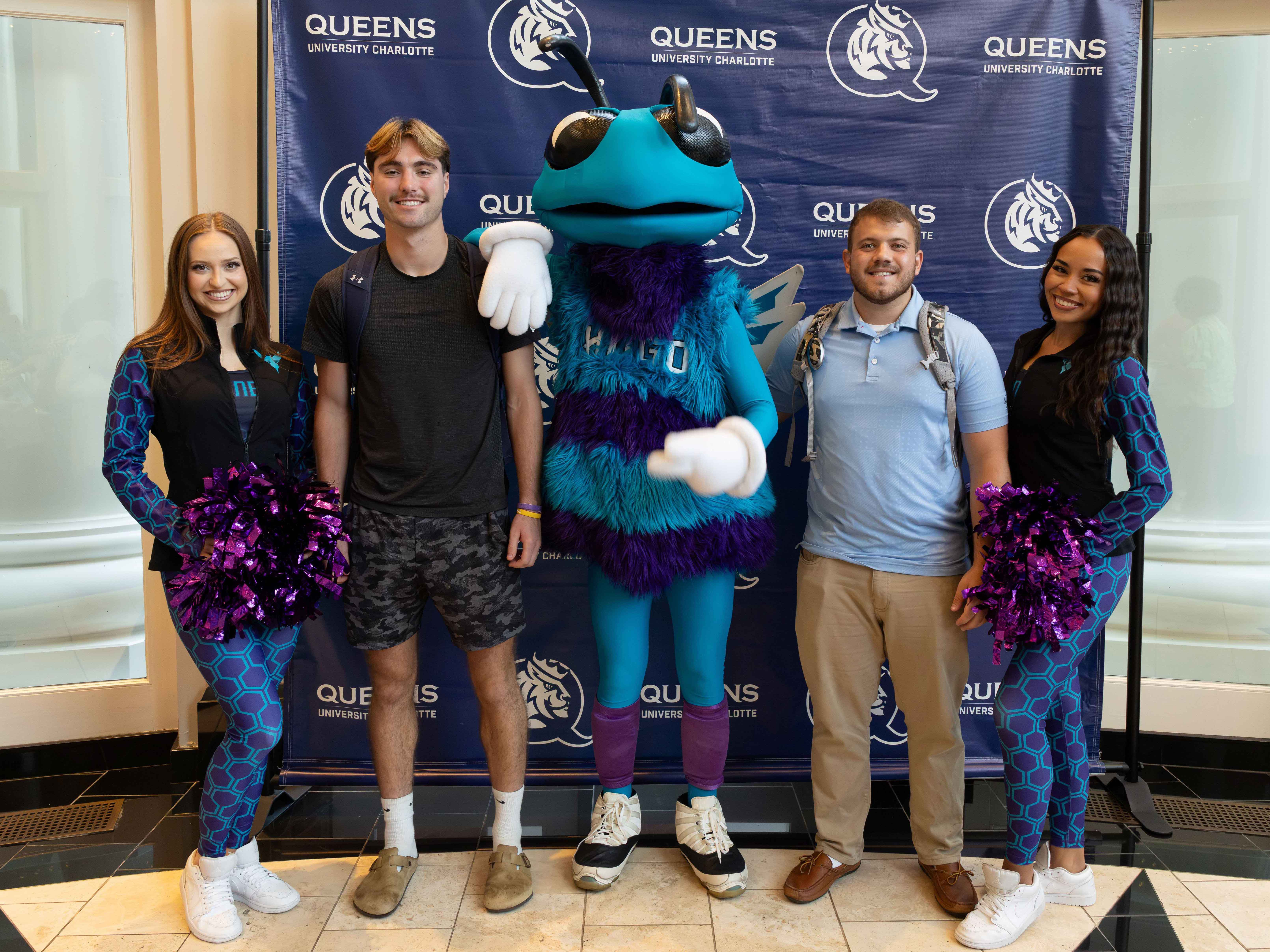 Two female dancers in teal and purple leotards flock male students with blue and teal bee Charlotte Hornets Mascot, mascot, Hugo.
