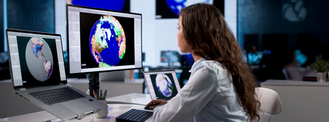 A woman in an office studies climate data maps of Earth on two large monitors, analyzing colorful weather patterns and geographic information, with a laptop and another screen nearby.