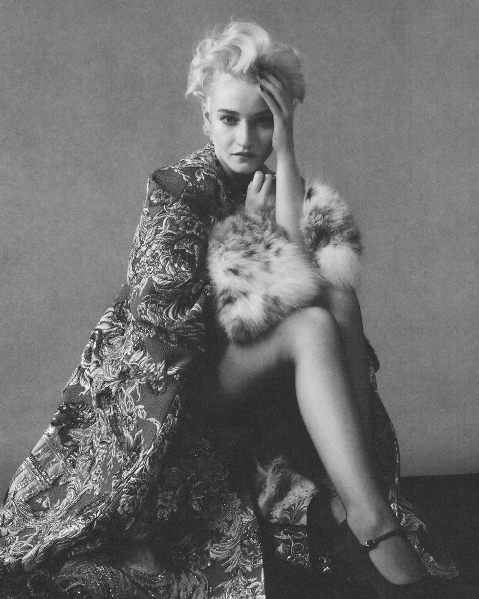 Studio portrait of Julia Garner in an ornate coat, resting her head on her hand, with a stylish fur accents.