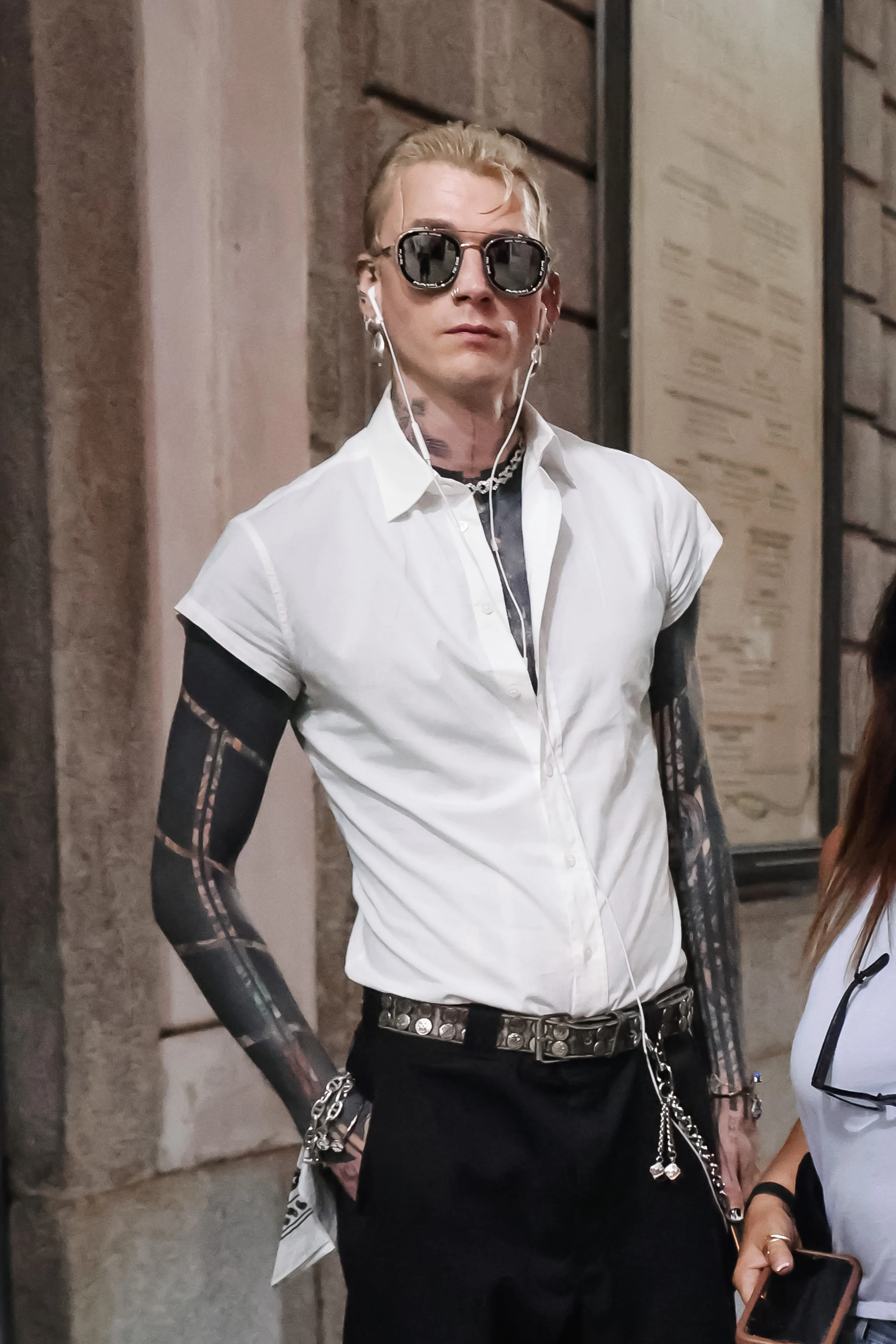 Machine Gun Kelly wearing a white short-sleeved button-up shirt, black pants, studded belt, sunglasses, and earphones during Milan Fashion Week.