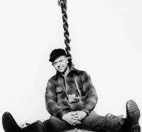 A man sitting on the ground with legs stretched out, wearing a black hat and a plaid shirt, with a large chain hanging above him.