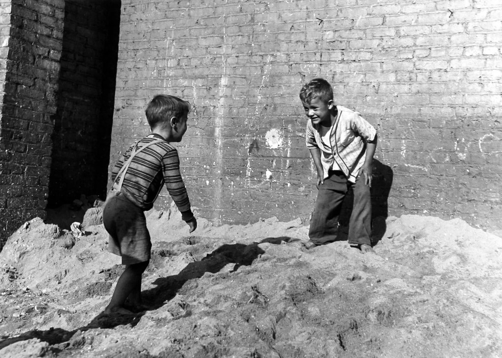 Two young boys playing in a sandy area, with one boy facing the other and smiling, while the other boy looks back at him. The background features a brick wall.