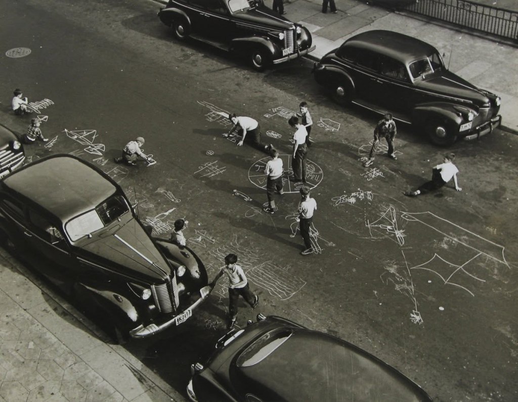 Children playing on a city street, drawing with chalk on the pavement near parked vintage cars.