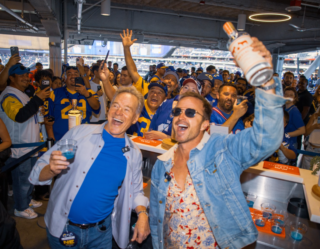 Two men drinking and holding a bottle of alcohol, smiling and cheering with a crowd at a stadium event.