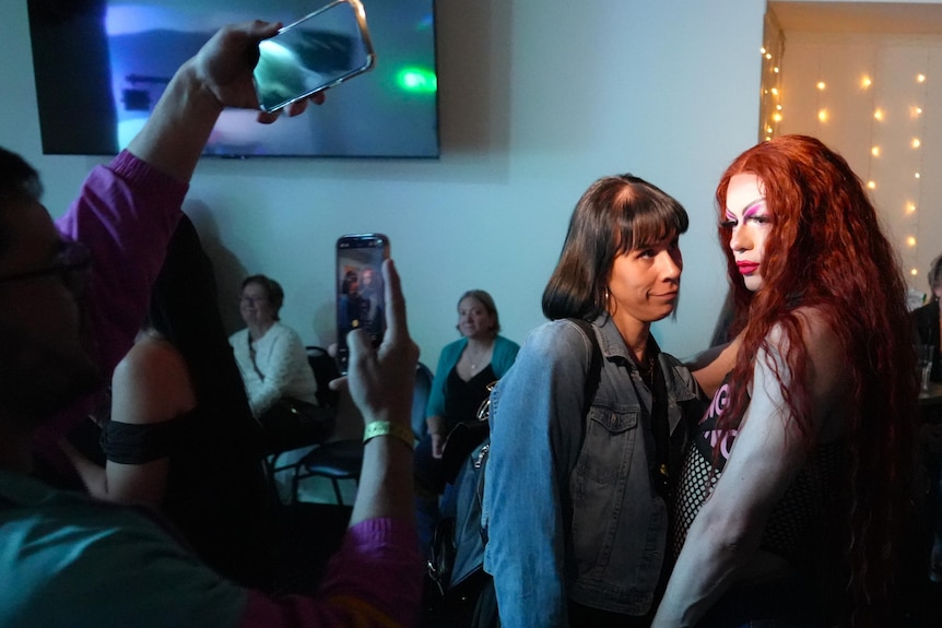A drag queen in a red wig poses for a photo with a brunette woman, surrounded by phones and other women.