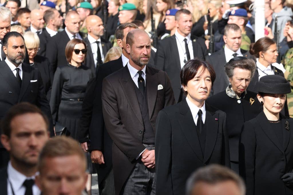 Prince William stands among a crowd in formal attire, looking solemn.