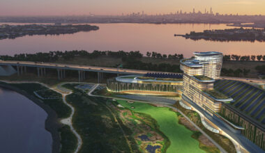 Modern waterfront building with curved architecture, surrounded by greenery and water, at dusk with a city skyline in the background.