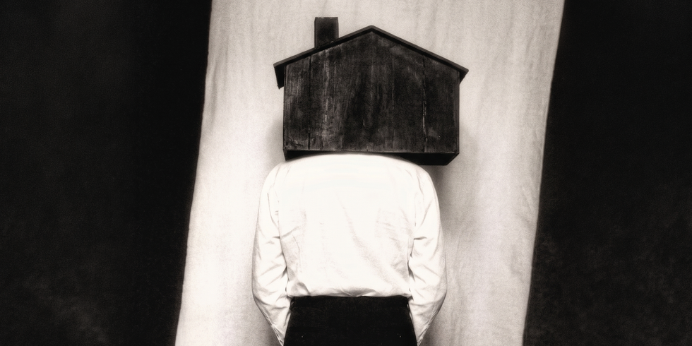 A person wearing a house-shaped object on their head, standing against a contrasting background.