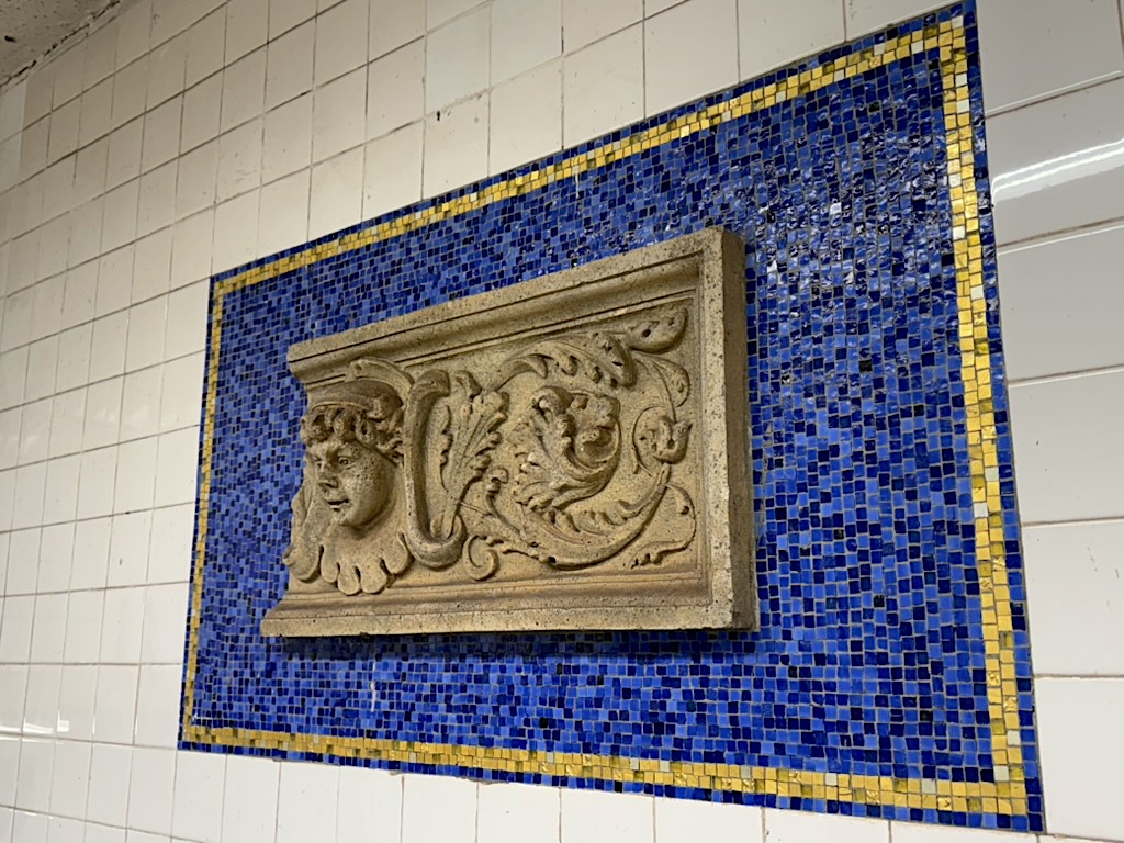 Relief panel featuring a face and a lion, set against a blue and yellow mosaic background in a tiled environment.