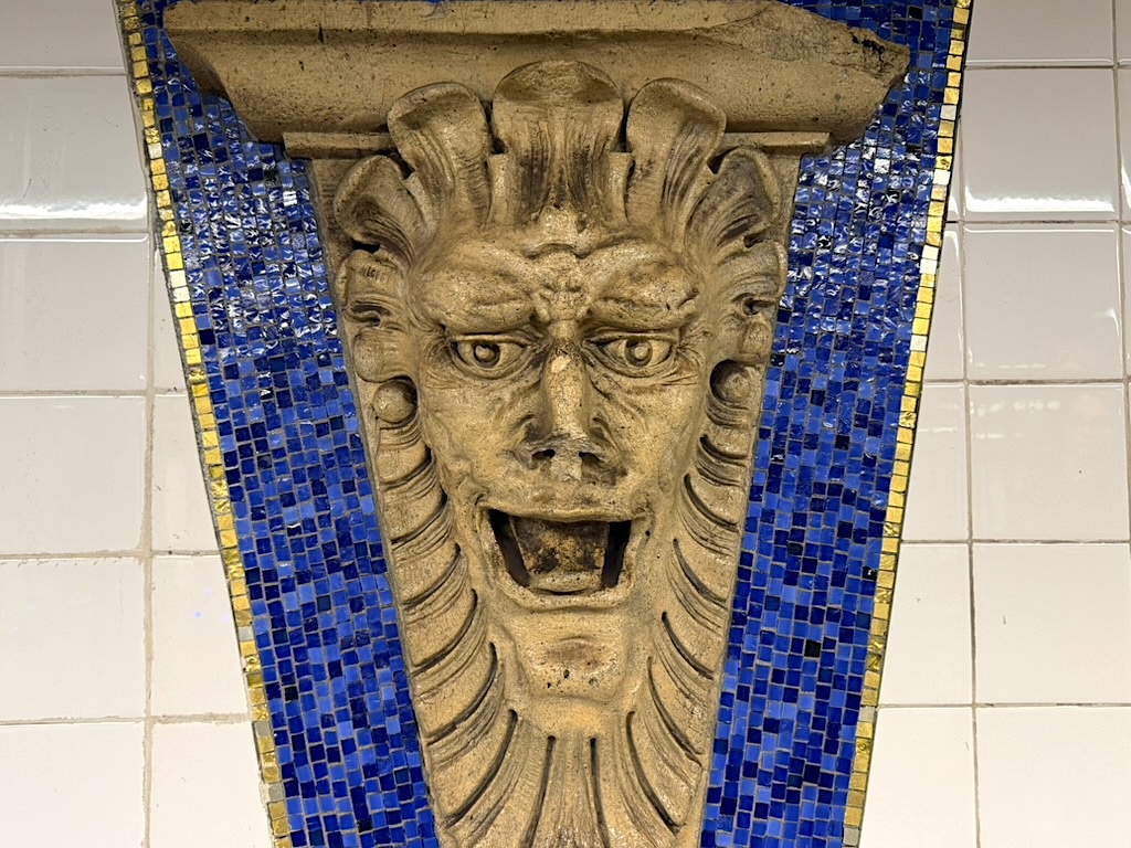Stone lion head sculpture with an expressive face, surrounded by blue and gold mosaic tiles, mounted on a wall.