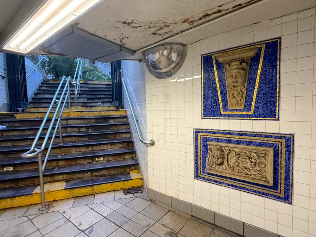 Interior view of a subway station showing stairs leading up with handrails, accompanied by decorative mosaic tiles featuring classical designs on the wall.