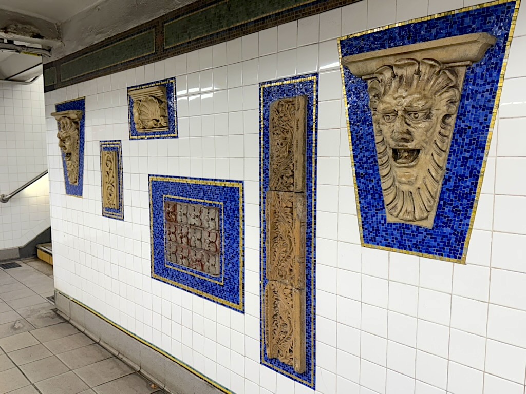 Decorative wall in a subway station featuring blue mosaics and sculpted lion heads among white tiles.