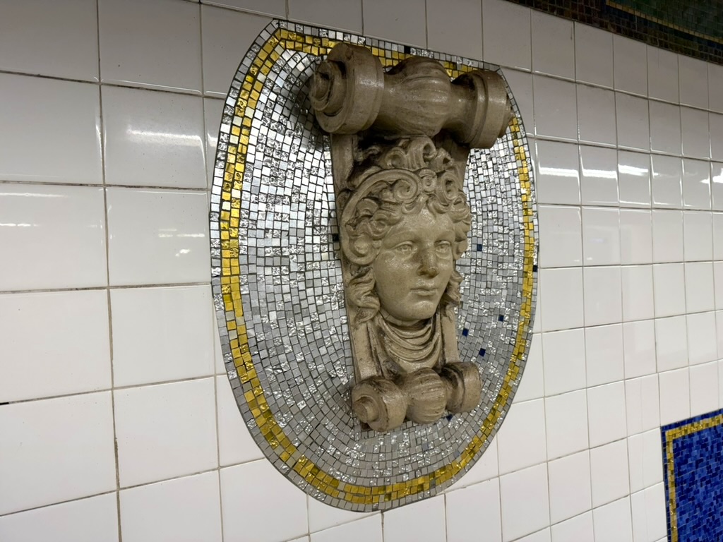 A decorative mosaic featuring a carved head with curly hair, set into a tiled wall in a subway station, surrounded by a patterned border.
