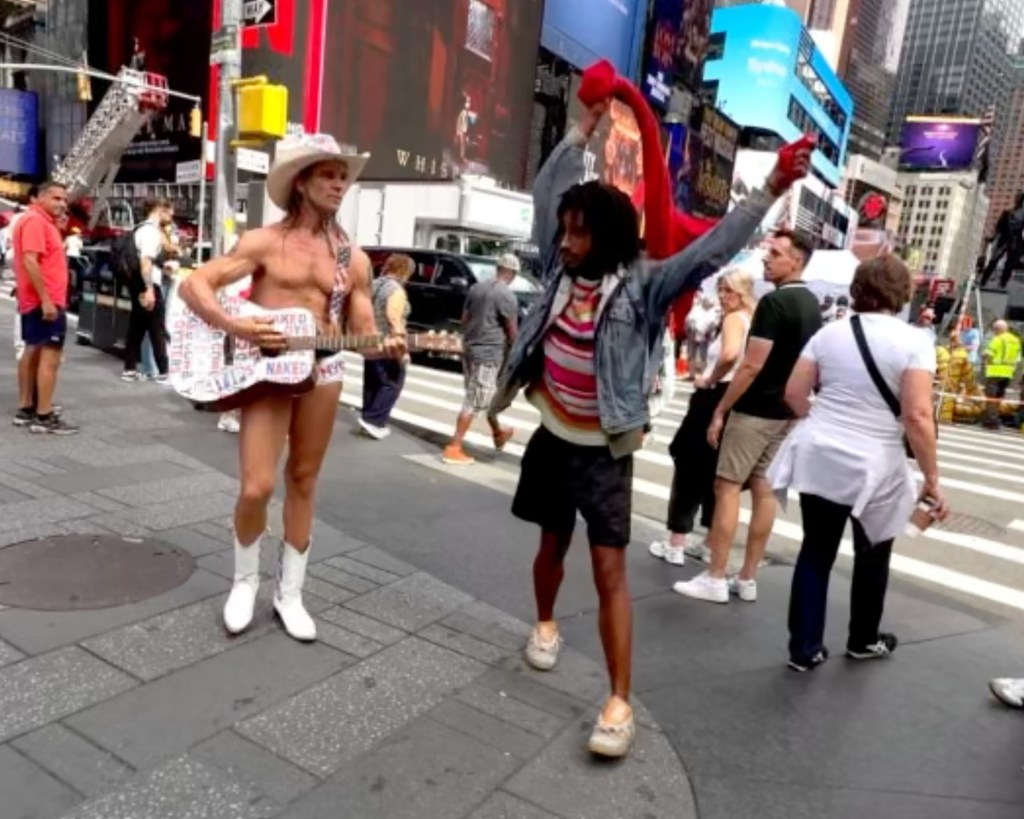 A bystander films the Naked Cowboy, wearing only a hat, boots, and briefs, with a guitar, as another man in a jean jacket and shorts, holding a red item, appears to harass him.
