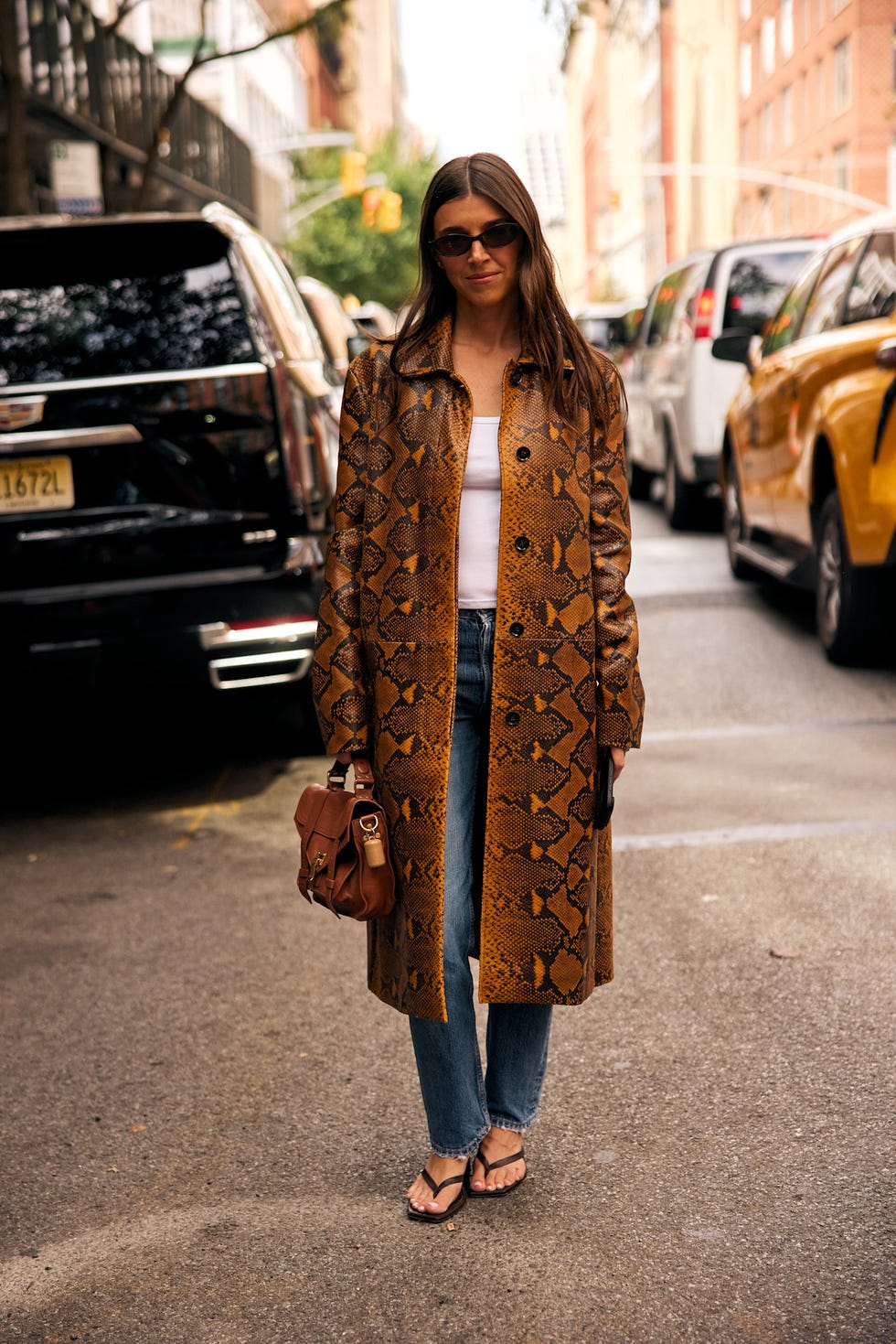 Woman wearing a snake-print coat, jeans, and sunglasses, holding a brown bag while standing on a city street. Woman wearing a snake-print coat, jeans, and sunglasses, holding a brown bag while standing on a city street.