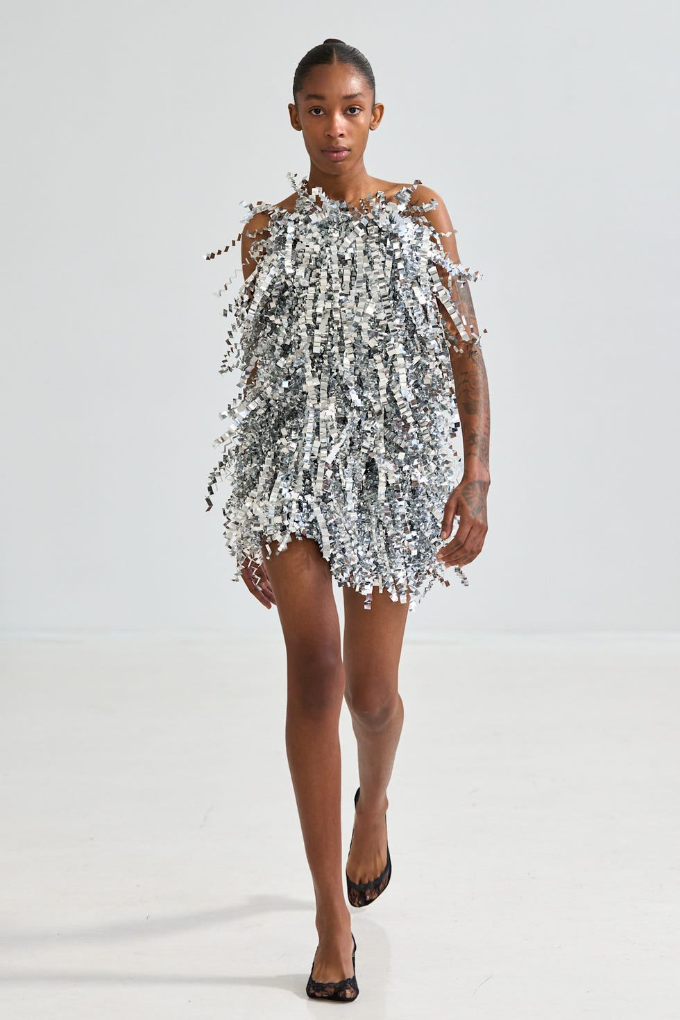 Model walking in a shiny, fringed silver dress on a minimalist background.