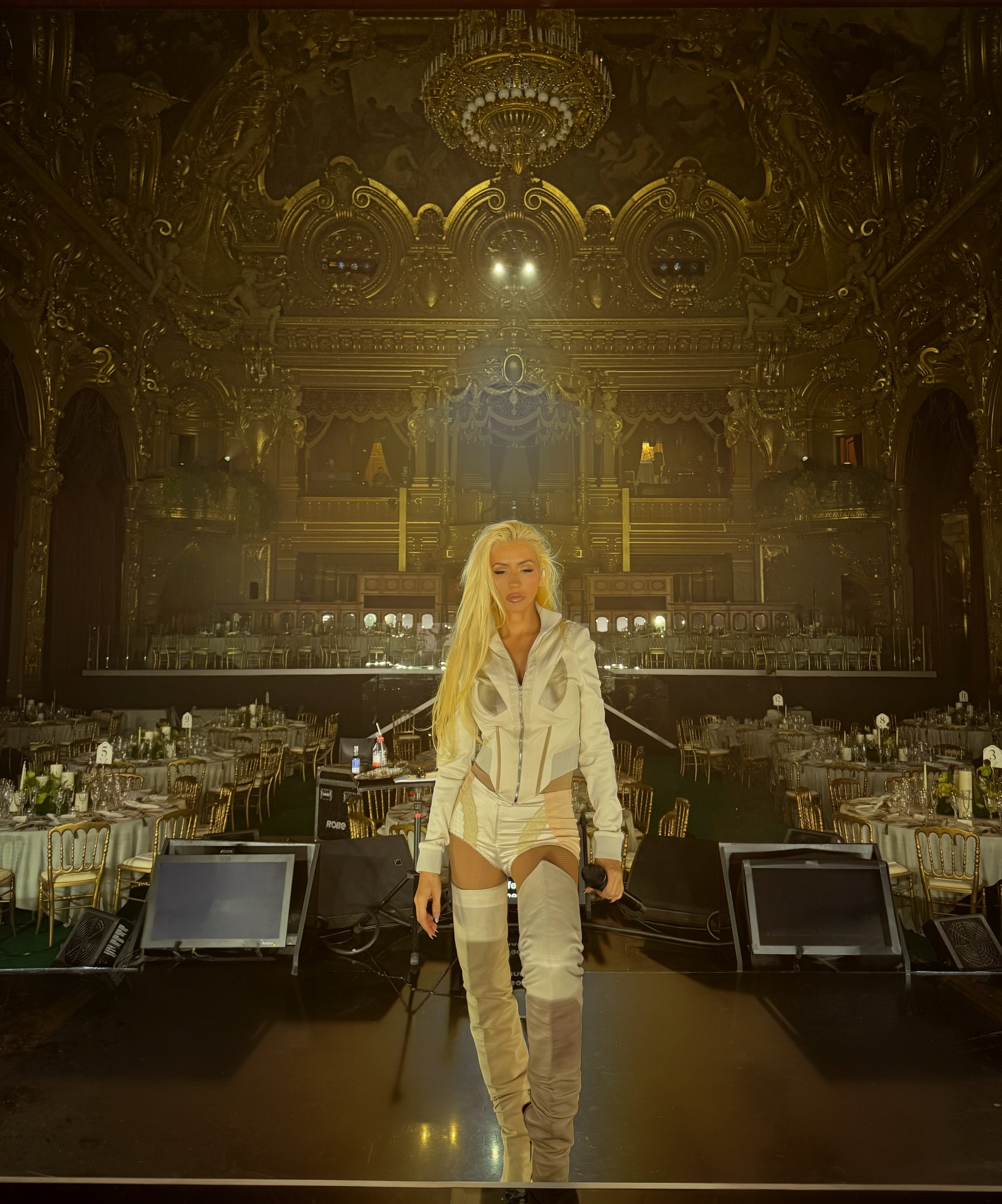 Christina Aguilera on a stage holding a microphone in a grand hall.