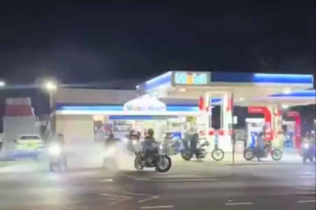 Motorcyclists gathered at a gas station, with smoke from burning tires filling the air.