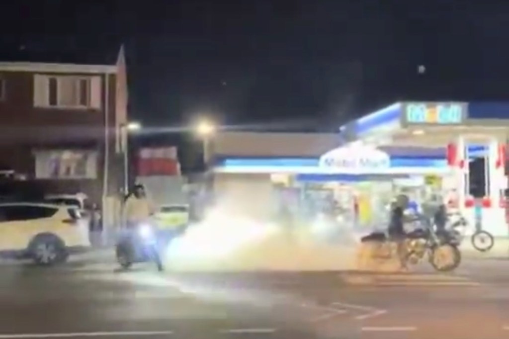 Police breaking up a street takeover, with a gas station in the background and smoke rising from a motorcycle's tires.